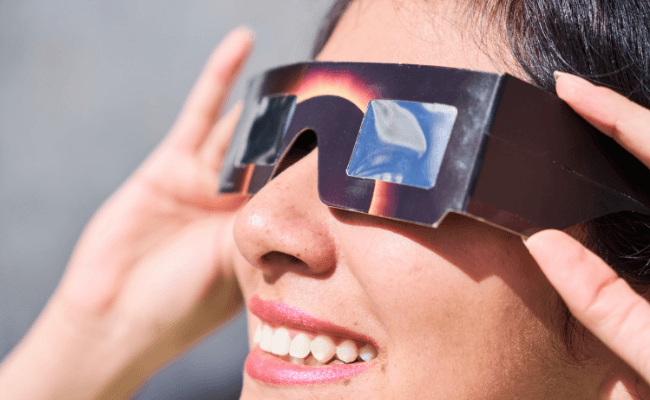 - Tracy Quinlan woman's face close up right profile shot. she's wearing eclipse glasses and looking up to a bright sky.